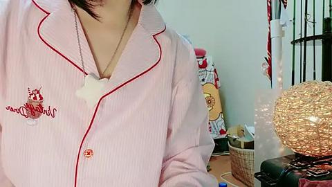 Media: Video of a person wearing a pink, striped pajama shirt with red piping and a dog tag necklace, indoors with a wicker basket, red toy, and a Christmas tree in the background.