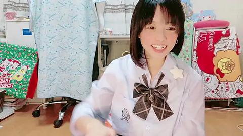 Media: Video of a cheerful Asian woman with straight black hair, wearing a light blue plaid shirt with a large bow tie, sitting in a cluttered office filled with colorful, patterned fabric pieces.