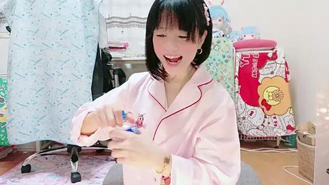 Media: Video of an Asian woman with straight black hair, wearing a pink pajama set, playing with a doll in a cluttered room with various colorful clothes and a dollhouse.