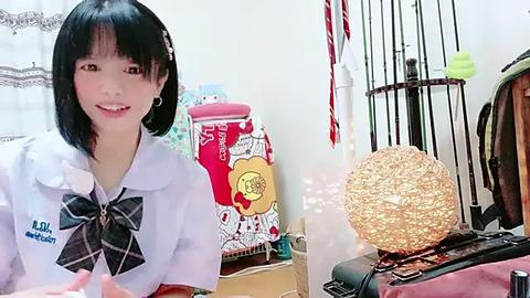 Media: Video of a smiling Asian girl with short black hair, wearing a white blouse with a plaid bow, in a cluttered room with a colorful cereal box, a round lamp, and a black metal cage.