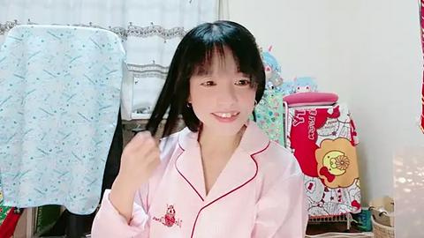 Media: Video of an Asian woman with long black hair, wearing a pink pajama shirt, smiling while brushing her hair, in a cluttered bedroom with white curtains, colorful quilts, and a red laundry basket.