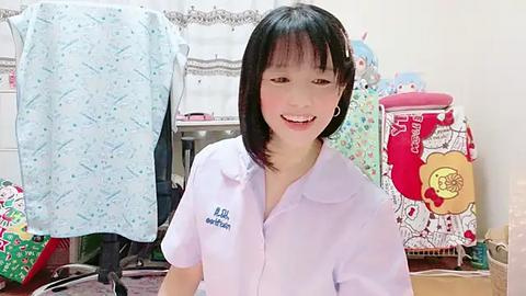 Media: Video of a young Asian woman with short black hair and a smile, wearing a light blue shirt, standing in a cluttered, colorful bedroom with a white bed and various toys and clothes.