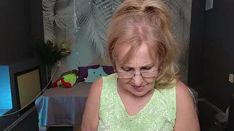 Media: Video of a middle-aged woman with short blonde hair, wearing glasses and a light green sleeveless top, sitting in a dimly lit room with tropical-themed decor.