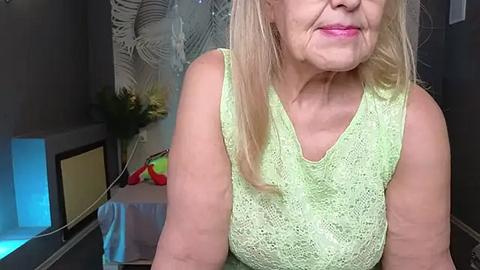 Media: Video of a middle-aged woman with light skin and shoulder-length blonde hair, wearing a light green lace sleeveless top, sitting in a dimly lit room with a blue-lit pool table and decorative wall art in the background.