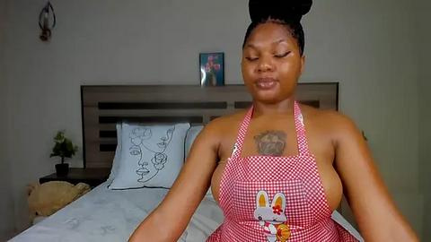 Media: Video of a Black woman with braided hair, wearing a red checkered apron with bunny design, sitting on a bed with white linens and floral pillow, in a simple, modern bedroom.