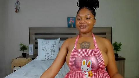 Media: Video of a smiling Black woman with braided hair in a red apron featuring a cartoon character, standing in a simple bedroom with a bed, plants, and abstract art on the wall.