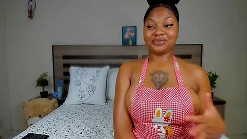 Media: Video of a smiling Black woman with medium skin tone and braided hair, wearing a red checkered apron with cartoon bunny design. She stands in a simple bedroom with a bed, a wooden headboard, and decorative pillows.