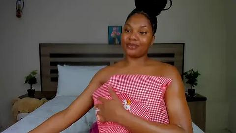 Media: Video of a smiling Black woman with a large bun in a red and white checkered dress, standing in a modern bedroom with a bed, nightstands, and a framed picture on the wall.