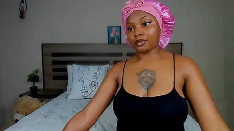 Media: Video of a dark-skinned woman with a pink headscarf, wearing a black spaghetti strap top, with a tattoo on her chest, in a modern bedroom with a gray headboard and floral art.