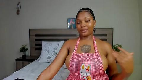 Media: Video of a Black woman with braided hair, wearing a red checkered apron with a bunny motif, standing in a modern bedroom with a white bed and minimalistic decor.