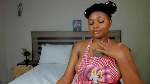 Media: Video of a smiling Black woman with short black hair in buns, wearing a red and white checkered apron, sitting on a bed with white sheets, in a simple bedroom with a wooden headboard.