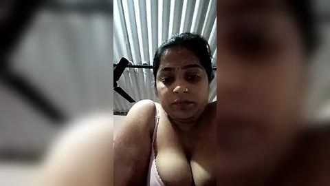 Media: Video of a middle-aged South Asian woman with dark hair, wearing a low-cut pink top, standing in a dimly lit room with a fan and vertical blinds.