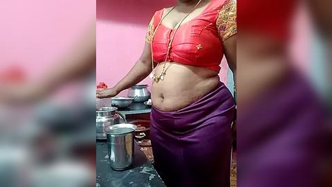 Media: Video of a woman with medium brown skin, wearing a red, ornate blouse and purple skirt, standing in a kitchen with pink walls, holding a ladle over a metallic pot.