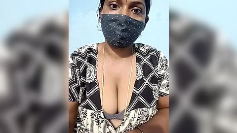 Media: Video of a South Asian woman with a medium brown complexion, wearing a black and white patterned blouse exposing cleavage, blue face mask, and a blue bra, against a blurred, grainy background.