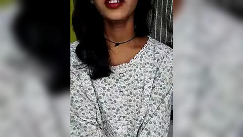 Media: A close-up video of a South Asian woman with medium brown skin, wearing a floral-patterned long-sleeve top, red lipstick, and a black choker necklace. The blurred background suggests an outdoor setting.