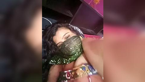 Media: Video of a woman with dark curly hair, wearing a green mask, lying on a pink couch, with a blurred background.