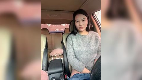 Media: Video of a young Asian woman with long black hair, wearing a gray knit sweater, sitting in the backseat of a car. The interior is beige with black seats and shopping bags visible.