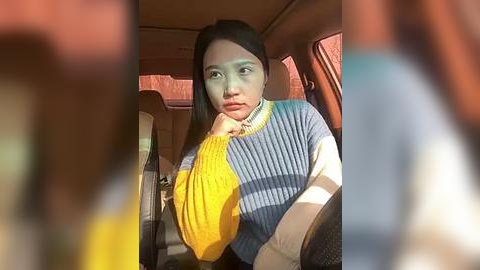 Media: Video of a young woman with long black hair, wearing a yellow and blue sweater, sitting in a car with blurred passengers.