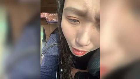 Media: Video of an Asian woman with fair skin and straight black hair, wearing a denim jacket, seated in a car. She has a contemplative expression, and her mouth is slightly open. The background features blurred, neutral-toned car interior elements.