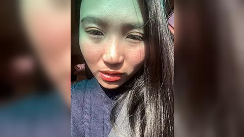 Media: Video of an East Asian woman with long black hair, light skin, and a neutral expression, wearing a dark blue sweater, taken in bright sunlight.