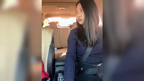 Media: Video of a young woman with straight black hair, wearing a navy blue sweater, seated in a beige leather car seat. She appears contemplative, with a slight frown, and a black crossbody bag slung over her shoulder. The car's interior is visible, with a red item on the seat beside her.