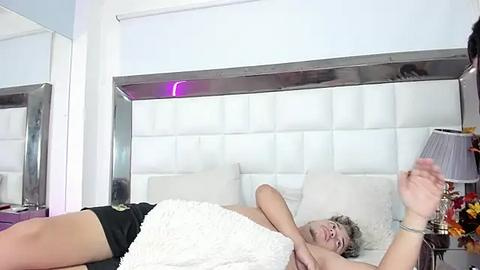 Media: A video of a woman with short, curly hair, wearing black shorts and a white blanket, lying on a white bed with a metallic headboard.