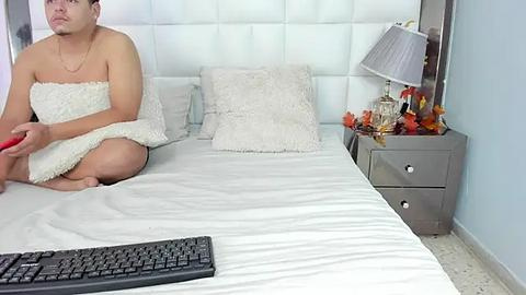 Media: Video of a man, nude except for a towel, sitting on a white bed with a white headboard, holding a red object, in a minimalist bedroom with a grey nightstand, lamp, and orange flowers.