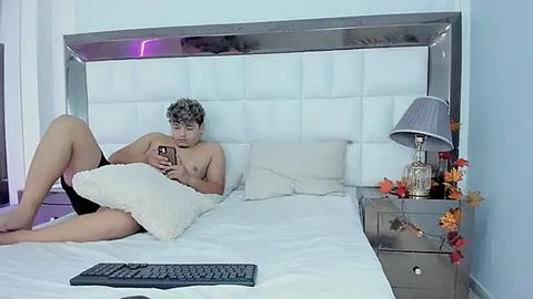 Media: Video of a young man with curly hair, shirtless, lying on a white bed with a white headboard, holding a smartphone, in a modern bedroom with a gray lamp, orange flowers, and a keyboard on the bed.