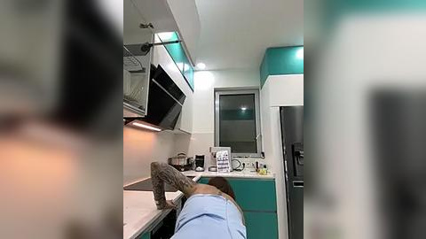 Media: Video of a modern kitchen with a person wearing a light blue shirt, leaning over a counter with a white oven above, and teal cabinets on the right.