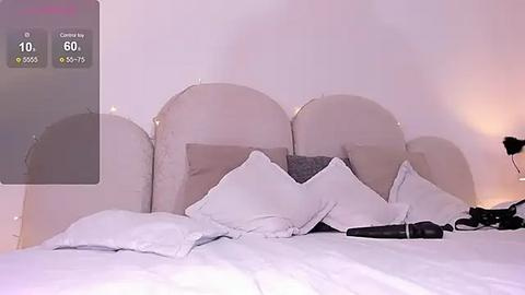 Media: Video of a modern bedroom with a white bed featuring three large, white pillows, a black remote control, and a smartphone displaying a camera app on the left.