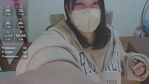 Media: Video of an East Asian woman with medium skin tone, wearing a beige mask, hoodie, and holding a phone in a dimly lit room.