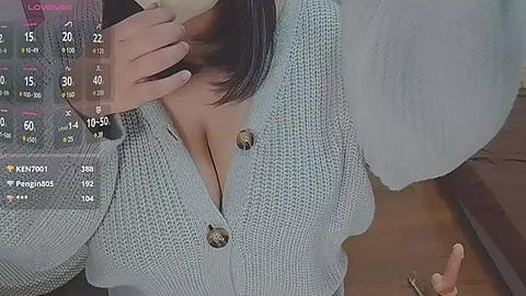 Media: Video of a woman with fair skin, wearing a light blue knitted cardigan with large buttons, holding her phone, and sitting on a bed with a white comforter.