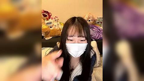 Media: Video of an East Asian woman with long black hair, wearing a white mask, sitting on a bed with plush toys and a purple blanket.