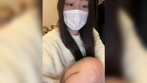 Media: Video of a young Asian woman with long black hair, wearing a white face mask and beige bathrobe, sitting on a bed with blurred background.