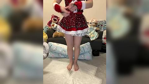 Media: Video of a person in a red, frilly, Santa-themed dress, barefoot, with Santa hat and gloves, dancing in a cozy, cluttered living room with plush toys.