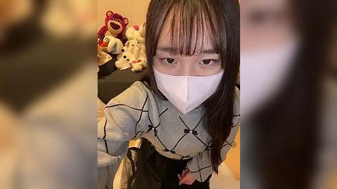 Media: Video of an East Asian woman with long, straight black hair and a white face mask, wearing a gray sweater with a geometric pattern, standing in a room with plush toys and a blurry figure.