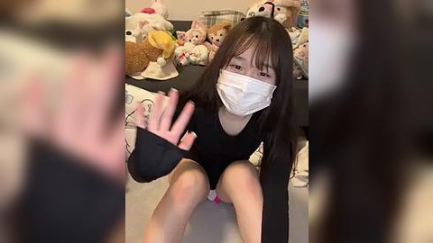 Media: A video of an East Asian woman with long black hair, wearing a black mask and black long-sleeve shirt, seated on a floor filled with stuffed animals. She gestures with her hand.