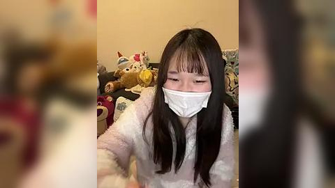 Media: Video of an Asian woman with long black hair, wearing a white face mask, pink pajamas, and a stuffed animal, seated on a bed with toys, in a dimly lit room.