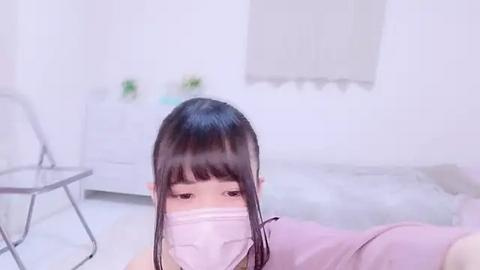 Media: A video of a young woman with long black hair, wearing a pink mask, lying on a bed in a bright, minimalist room with white walls and a white chair.