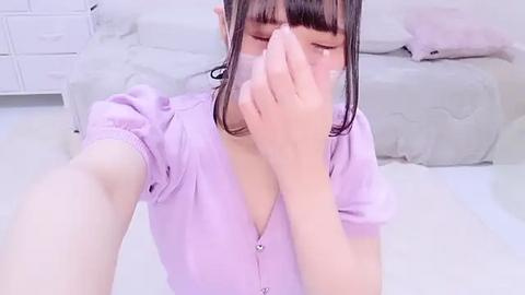 Media: Video of an Asian woman with straight black hair, wearing a light purple blouse, sitting on a white bed, covering her face with her hand, in a bright, minimalistic bedroom.