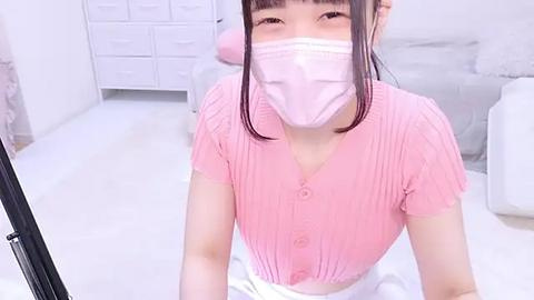 Media: A video of an Asian woman with light skin and long black hair, wearing a pink ribbed sweater, face mask, and white skirt, kneeling on a white floor in a minimalist, bright room with white furniture.