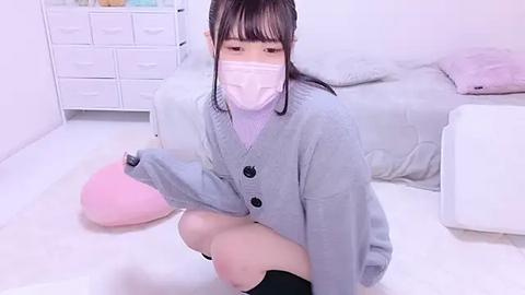 Media: Video of an East Asian woman in a pastel-pink face mask, light gray cardigan, and black shorts, kneeling on a white floor in a minimalist, white room with a bed and dresser.