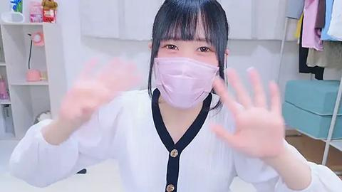 Media: Video of an Asian woman in a white blouse with a pink mask, smiling with hands raised in a clean, brightly lit room.