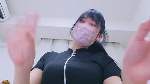 Media: Video of an East Asian woman with straight black hair and light skin, wearing a black zip-up hoodie, a purple mask, and a white shirt, holding up a pink object in both hands, indoors with a white ceiling.