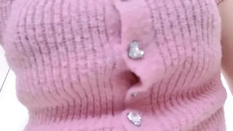 Media: Video of a fair-skinned person wearing a pink ribbed cardigan with two heart-shaped nipple piercings, one on each breast. The background is blurred, focusing on the intimate detail of the pierced nipples.