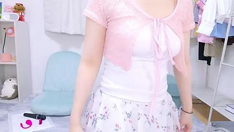 Media: Video of a slender woman with fair skin, wearing a pastel pink lace cardigan over a white tank top with a floral skirt. Background features a light blue chair, a white wardrobe, and a stuffed bear.