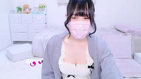 Media: Video of a young Asian woman with fair skin and dark hair, wearing a pink mask and a white ruffled dress with a black bow, standing in a minimalist, white bedroom with a bed, dresser, and stuffed animals.