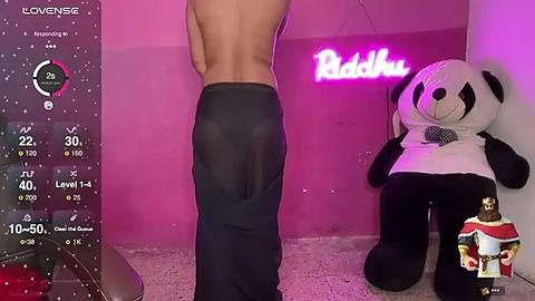 Media: Video of a topless man in sheer black pants, standing in a pink-lit room with a giant panda plush toy and a \"Buddha\" sign on the wall.