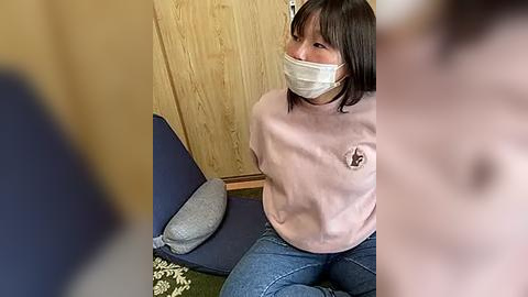 Media: Video of a young Asian woman with shoulder-length black hair, wearing a white surgical mask, pink sweatshirt, and blue jeans, sitting on a blue cushioned chair with wooden paneling background.