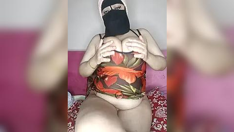 Media: Video of a light-skinned person wearing a black mask, floral-patterned bra, and panties, seated on a pink couch. Background is a plain white wall.